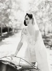 Black-and-white portrait of a bride in a lace gown and white gloves resting her hand on a wire garden chair, an old-world moment from a Santa Barbara wedding editorial by Yuliya Supin Design