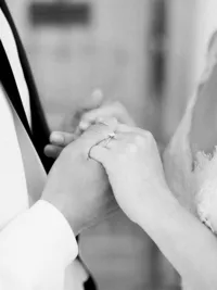 Close-up of bride and groom holding hands with wedding rings during Sunstone Winery ceremony.