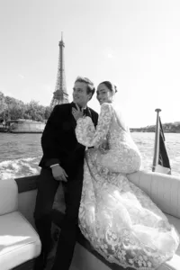 Black and white portrait of the bride and groom on the boat with the Eiffel Tower behind them, the bride in a long-sleeve lace gown