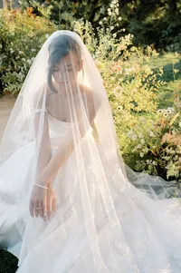 Bride seated quietly beside ceremony florals with veil softly covering her face in an intimate pre-ceremony moment.