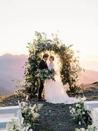 Bride and groom beneath a lush floral ceremony arch at sunset overlooking the mountains
