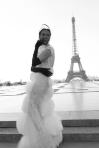 Black and white fashion portrait of a bride standing near the Eiffel Tower in Paris wearing a couture gown, long gloves, and a polka-dot veil headpiece that softly conceals her face.