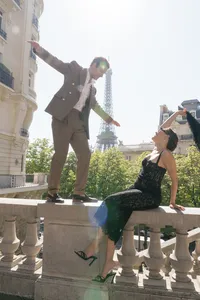 Bride sitting on a stone fence waving her scarf while the groom walks toward her along the fence with the Eiffel Tower visible behind them.