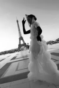 Black and white portrait of a bride walking toward the Eiffel Tower in Paris with a graceful hand gesture, wearing a couture gown and braided updo.
