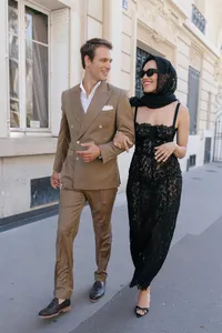 Bride and groom walking together through a Paris street during their engagement session, the bride wearing a black lace dress, silk scarf, and red lipstick while the groom wears a brown suit.