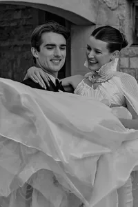 A joyful, fine-art black and white portrait of a newlywed couple embracing in front of historic stone architecture. The bride wears an elegant pearl necklace, and her voluminous gown creates beautiful, dramatic movement across the bottom of the frame.