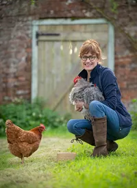 Environmental portrait of Jane Howorth, founder of the British Hen Welfare Trust, with a cockerel and hen in a garden setting.
