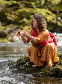 Virginia Compton kneeling beside the river, holding water in her hands during a photography session in Devon.