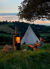 Rugby Player, Luke Arscott, relaxing beside a lit outdoor wood-burning stove, with a tipi and views across the Devon countryside.