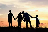 Professional photography by Nature's Child Photography: Palm Coast, family capturing a sunset silhouette of parents and their children standing hand in hand on grassy dunes, the little girl giving a thumbs-up against the orange horizon.