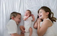 St. Johns newborn capturing a joyful sibling moment as a baby smiles between big brother and sister, their faces close and laughter shared in soft natural light. - A portrait experience by Nature's Child Photography.