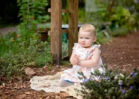 Baby girl in a floral dress sitting on a knit blanket among garden plants with a serious expression during an outdoor child in Mandarin. - A portrait experience by Nature's Child Photography.
