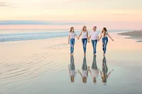 Professional photography by Nature's Child Photography: A family of four walks barefoot together at sunrise on Beach, their hands linked and reflections glowing in the shallow ocean water as they share a quiet morning moment during a fun family portrait session.