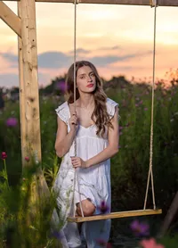Elkton, senior of a young woman in a flowing white dress holding a rustic swing at golden hour, wildflowers and pastel skies creating a dreamy, storybook feel. (Photographed by Nature's Child Photography)