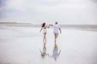 Professional photography by Nature's Child Photography: In St. Augustine, a couple runs barefoot through shallow water during a beach, wind tugging at their clothes as storm clouds gather in the distance.