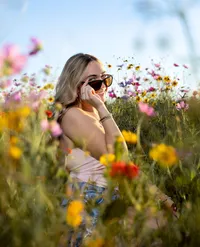 A young woman in sunglasses smiles while sitting among a colorful wildflower field, during her senior photo session. (Photographed by Nature's Child Photography)