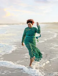 Jacksonville, FL portrait session capturing Beach, a grandmother in a flowing green lace dress kicks through the shoreline waves, laughing with carefree joy during a beach golden milestone session, a portrait session by Nature's Child Photography.