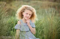 Young girl with curly blonde hair sitting in tall beach grass and smiling softly with her hand under her chin during a beach child portrait session. - A portrait experience by Nature's Child Photography.