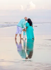 In Palm Coast, parents kiss gently on the beach while their toddler stands between them, turning to look at the camera as ocean waves and mirrored reflections stretch behind them. - A portrait experience by Nature's Child Photography.