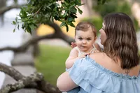 Mom and Me session featuring a mother holding her baby under a tree, the child’s bright blue eyes fixed forward with a bold, curious expression rather than a smile. - A portrait experience by Nature's Child Photography.