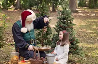 Young girl smiles as Santa shows her a handmade pinecone bird feeder in the Enchanted Forest, surrounded by decorated Christmas trees, dried oranges, woodland crafts, and the beauty of a Florida winter day.