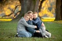 A mother kneels on green grass, smiling as her two young sons hug her from both sides. The boys lean in with big grins, their arms wrapped around her shoulders during a warm outdoor fall family session. (Photographed by Nature's Child Photography)