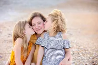 Two young sisters kiss their mother’s cheeks while sitting together on Beach, capturing a joyful and affectionate family moment by the ocean. - A portrait experience by Nature's Child Photography.