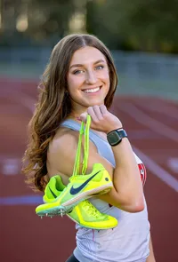 A graduating senior athlete smiles over her shoulder on a St. Johns, track, her bright racing spikes catching the light as she celebrates her final season. - A portrait experience by Nature's Child Photography.