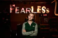 High school senior, stands with arms crossed under glowing “Fearless” signage, her steady expression adding attitude and strength to this urban senior sports session at a Jacksonville boxing gym. - A portrait experience by Nature's Child Photography.