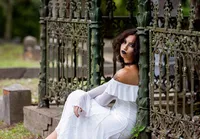Amelia Island portrait session capturing Senior in a flowing white dress sits against intricate wrought-iron gates in an cemetery, captured in a romantic, gothic-inspired senior. portrait session.