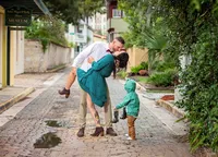 On a historic cobblestone road in St. Augustine, parents lean into a quick kiss as their little boy waits at their feet with his boots in hand, puddles reflecting the charm of a real family moment. (Photographed by Nature's Child Photography)