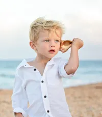Blonde haired boy holding a seashell up to his ear, listening to the oceans call in a child portrait session by Nature's Child Photography