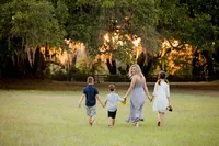 Professional photography by Nature's Child Photography: In Callahan, a mother leads her three children across an open field, looking back with a warm smile and sunflowers in her hand as they walk together beneath glowing oak trees during a family portrait session at a Callahan sunflower field.
