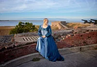 Senior of a high school graduate in a self-crafted historical dress, seated near a coastal fort with ocean views behind her. - A portrait experience by Nature's Child Photography.