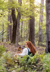 Jacksonville, FL portrait session capturing Orange Park, pet capturing a woman with long red hair sitting in a wooded area beside her tan dog, the dog seated upright and calmly watching the forest with her.