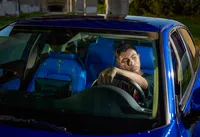 Photo of a young man resting his arm on the steering wheel of a blue sports car, framed in bold shadows and modern lines. - A senior portrait experience by Nature's Child Photography.