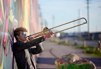 Professional photography by Nature's Child Photography: High school senior passionately playing trombone against a colorful industrial backdrop along a grafitti covered wall in St. Louis, Missouri, capturing movement, music, and confident individuality during his senior portrait session.