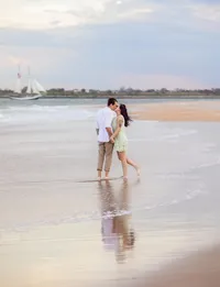 In Vilano Beach, a couple stands close in the shallow surf, foreheads nearly touching, their reflection mirrored beneath them while a sailboat glides across the horizon behind. - A portrait experience by Nature's Child Photography.