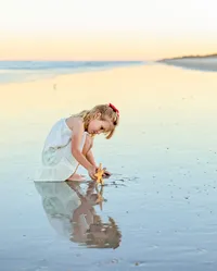 Young girl in a white dress kneeling in shallow water and examining a starfish as her reflection appears in the wet sand during a child portrait session on the beach. (Photographed by Nature's Child Photography)