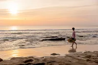 Professional photography by Nature's Child Photography: High school senior carries a wakeboard along the Palm Coast shoreline at sunrise, golden light reflecting across the ocean in a peaceful beach senior.