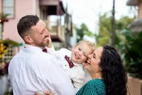 Professional photography by Nature's Child Photography: St. Augustine family capturing a mom and dad cuddling their son, his bow tie visible as he laughs in their arms against a softly blurred historic backdrop during a Nature's Child Photography Family Session.