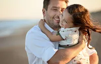 Professional photography by Nature's Child Photography: A joyful father cradles his daughter in his arms on the beach, her arms wrapped tightly around his neck as they share a warm, sunlit moment during a family photo session.