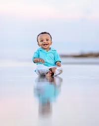 Professional photography by Nature's Child Photography: Laughing baby boy in a teal polo sitting barefoot on wet sand as his reflection mirrors him in the shallow water during a beach child photography session in Palm Coast.