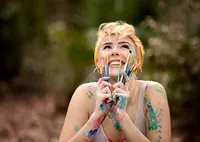 Artistic senior covered in bright paint grips a bundle of paintbrushes beneath her chin in Mandarin, Florida her joyful expression capturing bold creative spirit. (Photographed by Nature's Child Photography)
