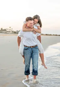 In Palm Coast, an engaged couple laughs during a beach as she wraps her arms around his shoulders and he carries her barefoot along the water during their engagement announcement. - A portrait experience by Nature's Child Photography.