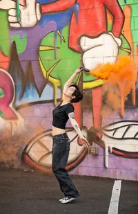 High school senior, holds an orange smoke flare aloft before a bold graffiti mural, creating a striking, high-energy senior portrait. (Photographed by Nature's Child Photography)