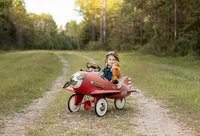 Professional photography by Nature's Child Photography: Baby wearing an aviator hat and overalls sitting in a red vintage pedal airplane and holding a stuffed teddy bear on a wooded trail during an outdoor child portrait session in St. Johns.