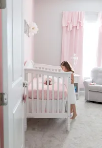 Professional photography by Nature's Child Photography: St. Johns newborn capturing a quiet sibling moment as a little girl gazes into the crib at her baby sister, surrounded by soft pink nursery details in their home.