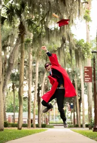 St. Augustine senior jumps high in graduation robes beneath moss-draped oaks at Florida School for the Deaf and Blind, celebrating commencement with movement and triumph in a dynamic senior session. (Photographed by Nature's Child Photography)