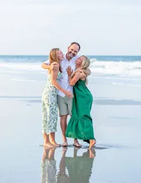 Jacksonville, FL portrait session with Nature's Child Photography capturing Beach family of a father holding his two daughters tight in the wet sand, their dresses brushing the shoreline as they smile up at him.
